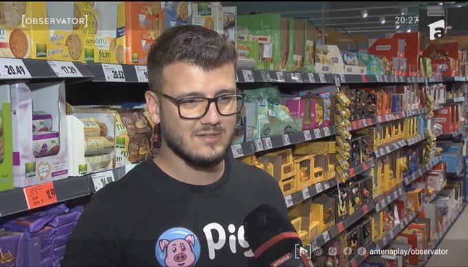 A young man with glasses being interviewed by AntenaPlay Observator in a supermarket aisle stocked with various packaged snacks and cookies priced in Romanian lei.