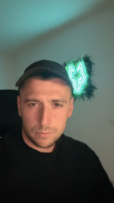 A man wearing a black cap and black shirt takes a selfie in a dimly lit room, with a glowing green neon wolf head sign mounted on the wall behind him.