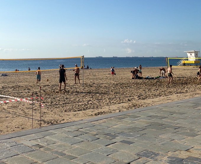 A beach scene with people playing volleyball on a yellow net court, a lifeguard tower on the right, and ships visible in the distance across the ocean.