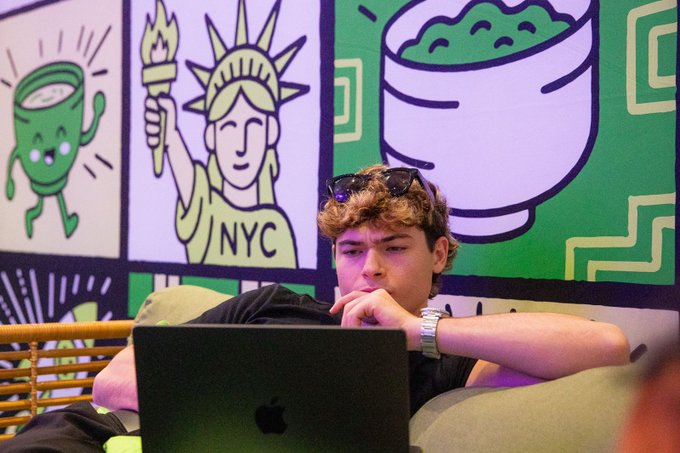 The image shows a young man sitting on a couch with an open laptop computer in front of him. The background features a green wall adorned with various cartoon characters, including a Statue of Liberty and a bowl of food. The text "As it turns out, Matcha Park is a great place to cowork from ETHConf" can be seen, suggesting that the man might have been working on a project related to cryptocurrency or technology.