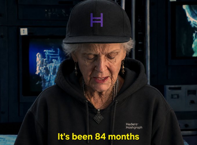 A meme image showing an elderly woman wearing a black Hedera Hashgraph cap and hoodie with the caption "It's been 84 months".