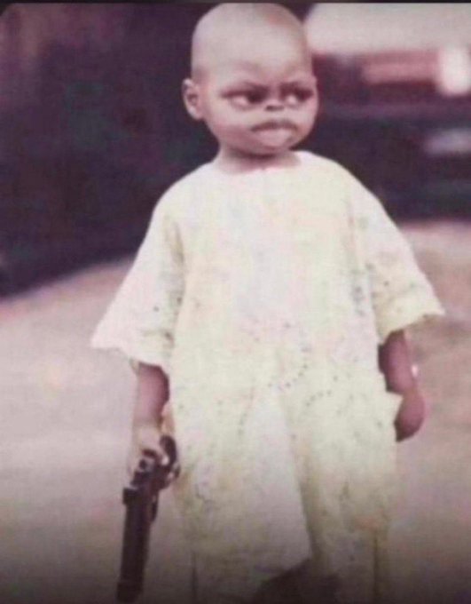 In the image, a young child dressed in a white dress stands next to a gun, with their face obscured by glasses. The child appears to be posing for the camera while holding the gun. The background of the photo shows a street scene, which adds context to the overall setting and atmosphere. The caption at the bottom of the image reads "Good morning web3 warriors Will you say it back Another day in the trenches with @useTria and @River4fun Got eyes on @TheARCTERMINAL too What are you focusing on this new month Happy new month to them all".
