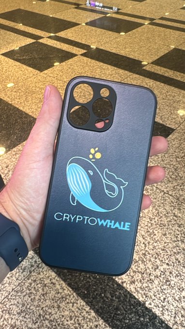 A hand holds a black smartphone case featuring a blue whale logo and the text 'CRYPTOWHALE' over a checkered floor.