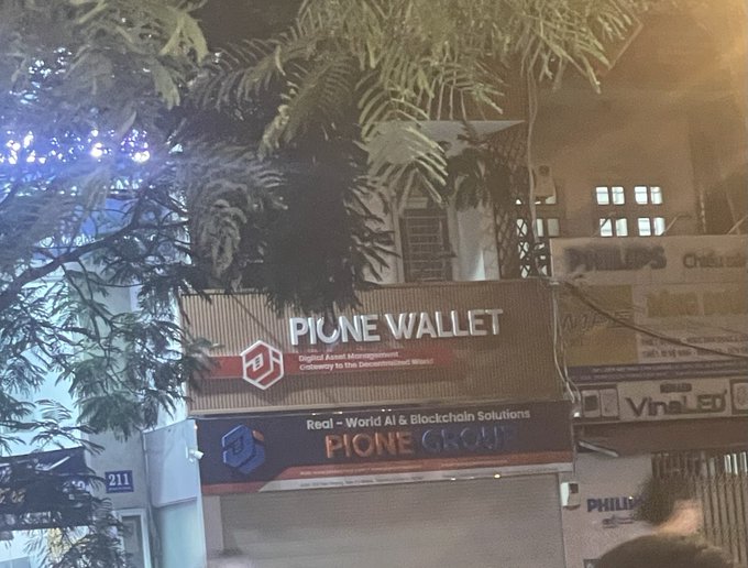 A nighttime street view of a storefront sign reading 'PIONE WALLET' with text below stating 'Real-World AI & Blockchain Solutions' and 'PIONE GROUP'.