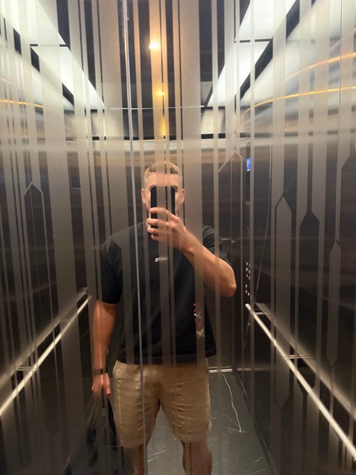 A mirror selfie of a man with braided hair wearing a black t-shirt and tan shorts, taking the photo inside an elevator with distinctive vertical striped glass panels.