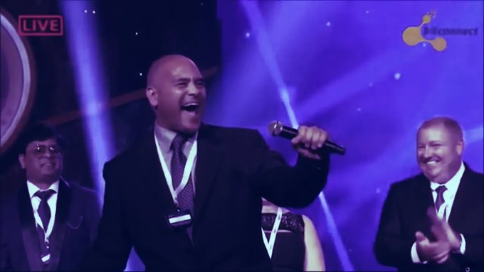 A bald man in a dark suit enthusiastically sings into a microphone on a purple-lit stage during a live broadcast, flanked by other formally dressed men including one who is clapping.