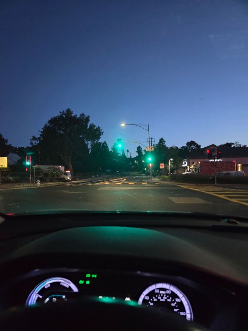 A first-person view from inside a car at dusk shows the illuminated dashboard and a road intersection with green traffic lights ahead, lined with trees and buildings under a deep blue evening sky.