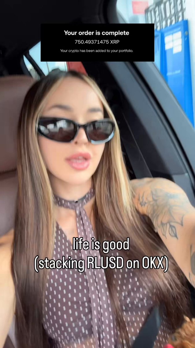 The image features a woman wearing sunglasses, with long hair, sitting inside a car. The background of the photo shows an advertisement for Ripple, which promotes its "Running RLDUSD" campaign. This campaign encourages users to deposit at least $6 into Ripple's cryptocurrency wallet and offers a 6% deposit match in RLUSD.