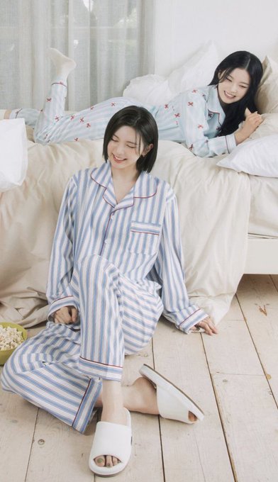 Two young women smile while wearing matching blue and white striped pajamas in a bright, cozy bedroom setting.