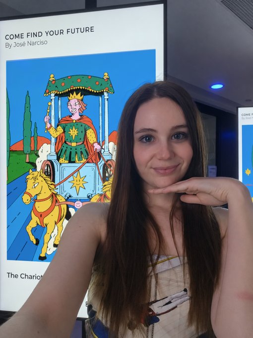A young woman with long brown hair poses next to a poster titled 'COME FIND YOUR FUTURE By José Narciso' featuring an illustration of 'The Chariot' tarot card with a crowned figure.