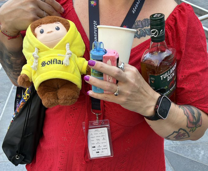 A person in a red dress holds a small brown stuffed monkey wearing a yellow 'golf' hoodie, while also carrying a bottle of 'TULLAMORE DEW' whiskey, a paper cup, and lighters.