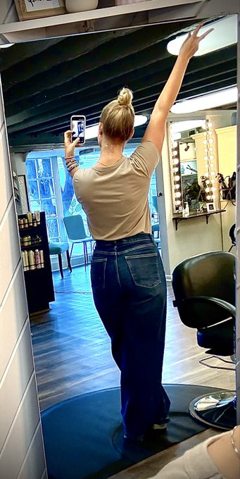 A blonde woman with her hair in a bun takes a mirror selfie in a salon, wearing a tan t-shirt and dark blue jeans.
