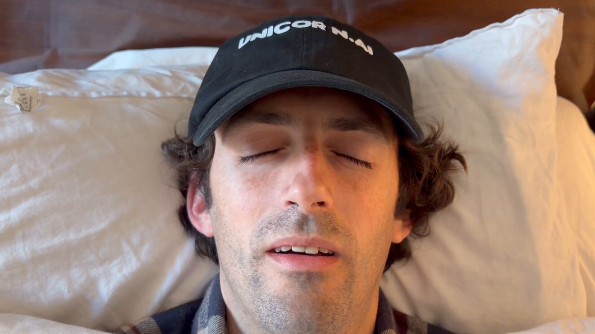 The image shows a man lying down with his eyes closed, wearing a black baseball cap that has " unicorn nam" written on it. There are two pillows visible behind him - one on each side of his head. The caption below the photo reads "Everyone needs to use Pancake right now".