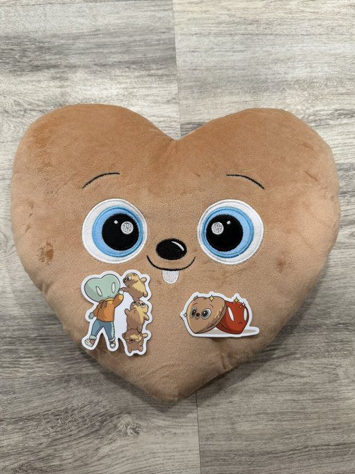 A plush brown heart-shaped pillow featuring a cute cartoon face with big blue eyes and a smile is displayed on a wooden floor. The pillow has two character patches on it: one shows a green-headed figure hugging a bear-like character (likely from LT3NFT), while the other features a brown furry creature with a red heart (possibly from FugzOfficial), tying directly to the post about receiving this comfy branded merchandise.