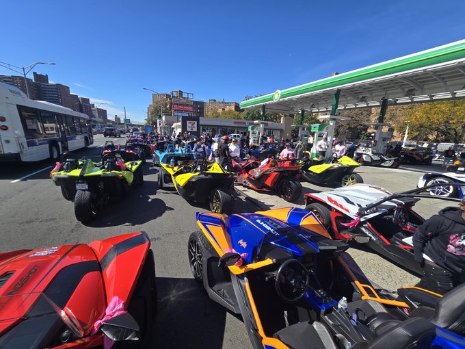 A large gathering of colorful Slingshot three-wheeled motorcycles and ATVs parked at a BP gas station, with a crowd of people and a bus visible in the background on a sunny day.