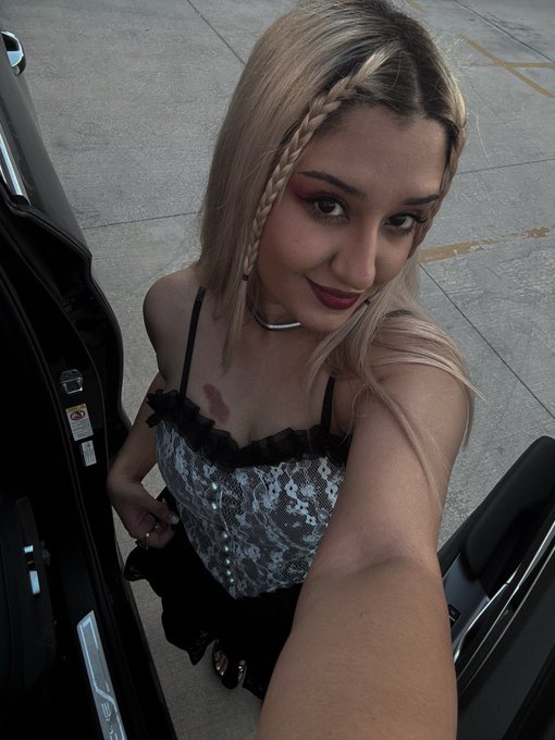 A woman with blonde braided hair wearing a black and silver sequined corset-style top takes a selfie inside a vehicle.