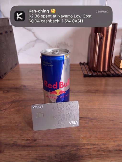 The image features a can of Red Bull soda sitting next to a credit card, suggesting that the person who took this photo is interested in purchasing the energy drink or has recently purchased one. The post includes information about KAST being a "bank card" offering similar benefits as those found with more traditional payment methods. These benefits include instant checkout at local shops and a 1.5% cashback reward for purchases made within their network, which can directly be deposited into the credit card's account once made. The text in the post emphasizes how this card works exactly like a regular bank card and is designed to make transactions easier and more convenient for users.