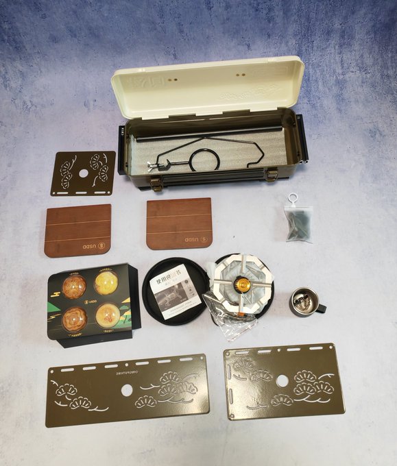 A collection of camping and cooking accessories is laid out on a surface, including a green metal stove/case, wooden trivets with the 'asan' logo, metal stencils, and small cooking pots.