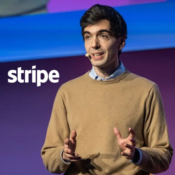 A man in a tan sweater speaks on stage with a headset microphone, gesturing with both hands while the "stripe" logo is visible in the background. The image accompanies news about Stripe's plans to expand stablecoin card programs to 100+ countries by end of 2026, suggesting the speaker is a Stripe executive announcing this payments infrastructure initiative.