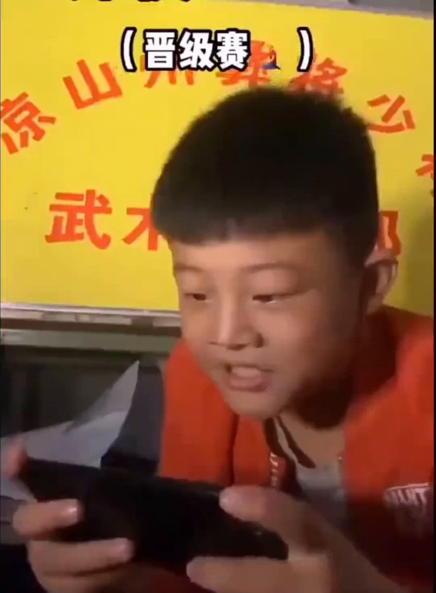 The image shows a young boy playing with a cell phone while wearing an orange shirt. The boy appears to be looking at something or someone on his phone screen, possibly reacting to a message or notification. The background of the image features a yellow sign with Chinese characters and some text in English.
