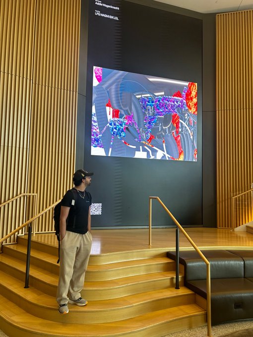 A person wearing a black shirt, tan pants, a backpack, and a VR headset stands on a wooden tiered platform looking at a large screen displaying a colorful virtual environment.
