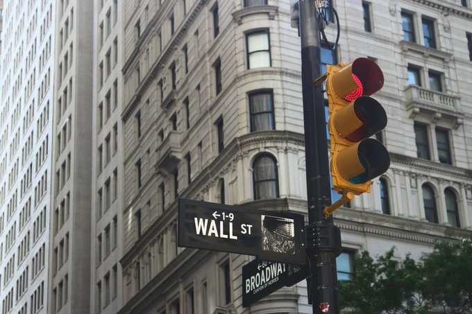 A street sign for Wall St and Broadway stands in front of a classic stone building, with a traffic light showing red and green.