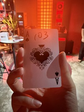 A hand holds an Ace of Spades playing card with '103' written on it and an ornate spade design featuring two figures, viewed in a dimly lit room with red accents.