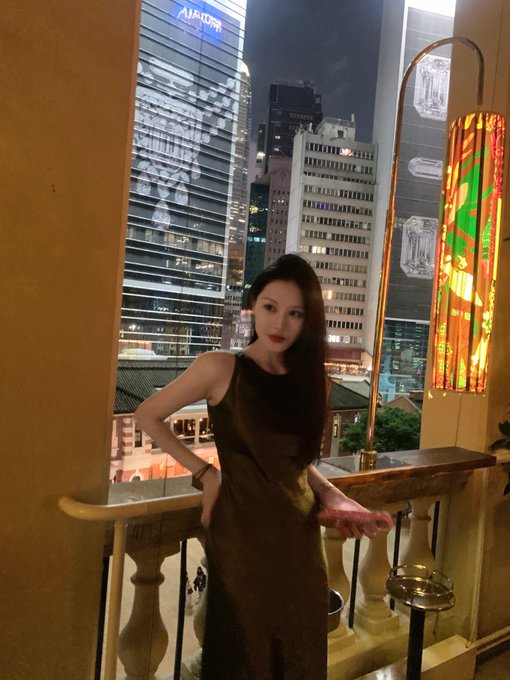 A woman with long dark hair wearing a sleeveless brown dress poses on a balcony at night, with an illuminated "AVALON" building and other city skyscrapers visible in the background.