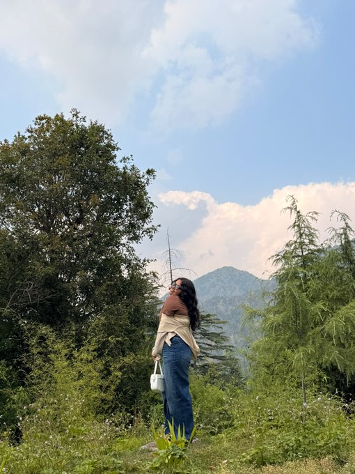 A young woman with long dark hair, wearing a brown top and jeans, gazes up at a partly cloudy sky surrounded by lush green trees and distant mountains, carrying a small white bag. The serene, expansive natural scene mirrors her caption's ambitious and grounded self-introduction—balancing her ambitions of reaching seven figures with the steady presence of her roots in nature and reality.