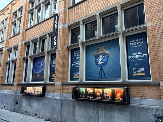 The image shows a brick building with large windows, advertising an event called "The Litecoin Summit". The advertisement features a cartoon character wearing a cowboy hat and holding a litecoin sign. There are also several movie posters displayed on the side of the building. A bicycle is parked in front of the building, suggesting that people might have ridden their bikes to attend the summit or simply pass by while walking down the street.

The post about the Litecoin Summit invites people to "get ready for the Litecoin Summit in Amsterdam!". The image also includes a reference to the "Litecoin Summit" and possibly some information about the event, such as its location (Amsterdam) and the time it is taking place (Saturday).