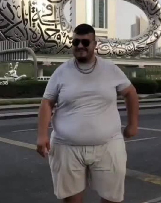 A man in a white t-shirt, beige shorts, and sunglasses walks confidently down a street in Dubai, with the Museum of the Future's Arabic calligraphy visible in the background. The caption "He is my bitch" humorously suggests the photo was taken as the subject jokingly approached the photographer.