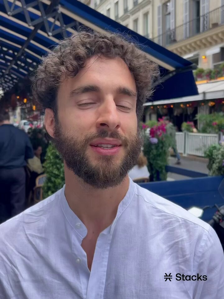 The image shows a man with curly hair wearing a white shirt, standing outside of a restaurant or cafe. The photo has been edited to include a watermark that reads "Stacks @ proofoftalk" above the image.
