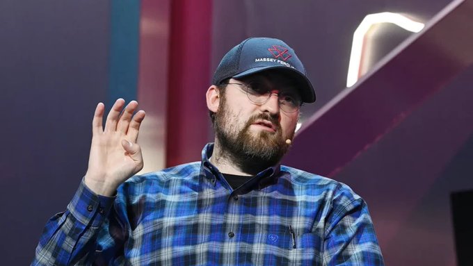 A bearded man wearing glasses, a blue plaid shirt, and a 'Hassee' branded cap, speaking and gesturing with his hand while wearing a headset microphone on a stage.