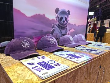 The image shows a display table with several purple hats arranged on top. The hats are positioned at different angles, showcasing their unique designs. In front of the hats, there are books that appear to be promotional materials for an event or exhibition. A person can also be seen in the background, possibly attending the event or browsing through the displayed items.