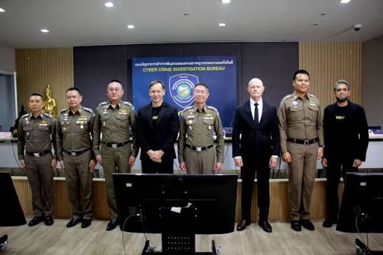 A group of eight officials, including Thai police officers in tan uniforms and two men in dark suits, pose for a photo in front of a screen displaying the "Cyber Crime Investigation Bureau" logo. The image relates to the post by illustrating a high-level law enforcement or government meeting, suggesting the involvement of Thailand's cyber authorities in investigating the alleged $307M/year USDT laundering case and the country's push toward a baht-backed stablecoin.