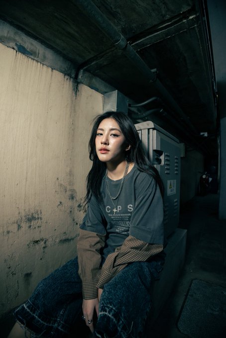 A young woman in a dark t-shirt, silver chain necklace, and baggy jeans sits in a dimly lit, gritty industrial corridor.