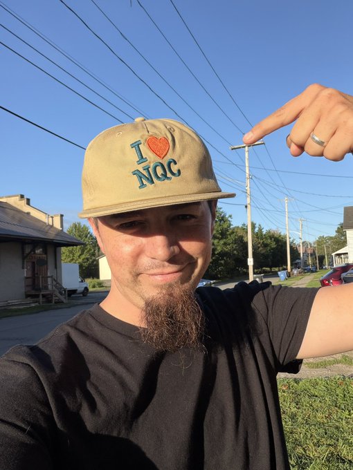 A man wearing a tan 'I ❤️ NQC' cap with a long goatee poses for a selfie on a sunny suburban street, pointing his finger toward the sky.