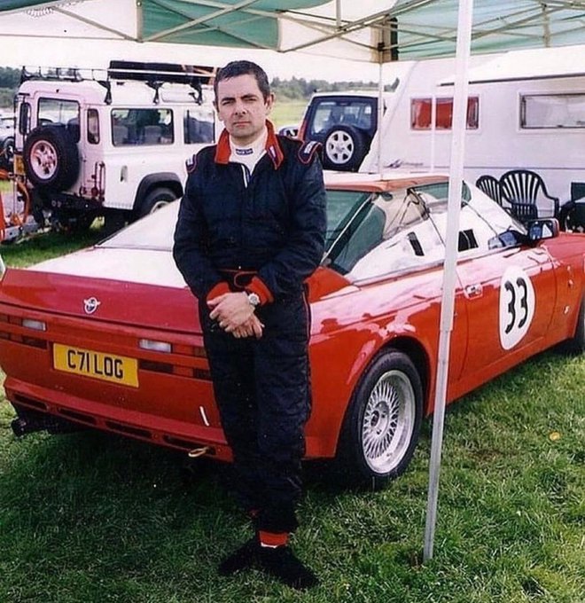 A man in a black racing suit stands in front of a red racing car with the number 33 on the side and license plate C71 LOG.