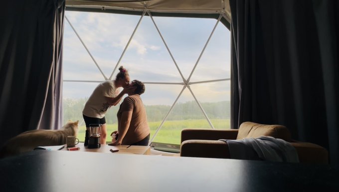A couple shares a tender kiss in front of a large geometric window overlooking a green landscape, with a French bulldog resting nearby, celebrating an engagement with a ring. The image captures an intimate, romantic moment that aligns with the post's "PUT A RING ON IT" announcement.
