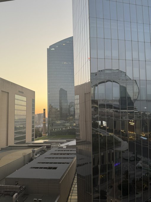 A view of modern glass skyscrapers at sunset, with one building's reflective facade showing a distorted image of another tower.