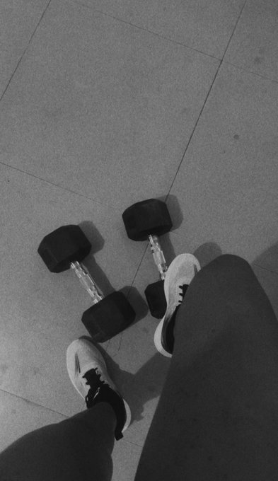 Black and white photo looking down at a person's feet in sneakers standing next to two dumbbells on a tiled floor.