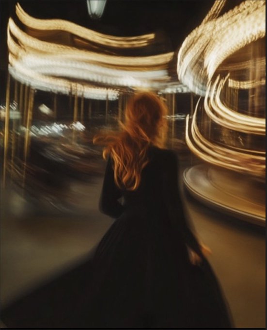 A blurred, long-exposure photograph of a woman with long red hair wearing a dark dress, standing in front of a spinning carousel illuminated with streaks of golden light.