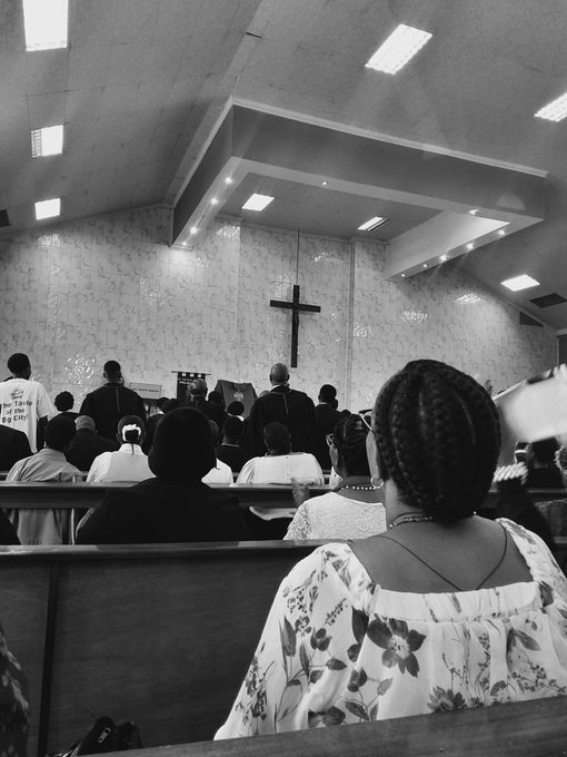A black and white photograph taken from behind congregants seated in wooden pews inside a church, with a large cross visible on the front wall and a woman with braided hair in the foreground.
