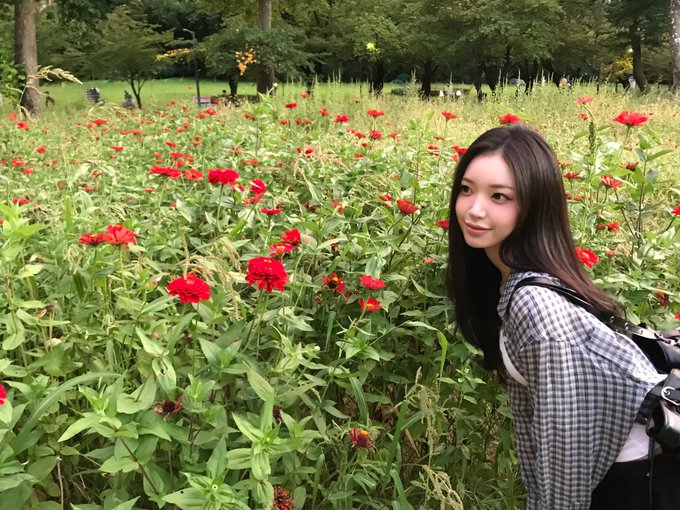 A young woman with long dark hair poses for a photo in a field of red zinnia flowers, wearing a checkered shirt and carrying a backpack.