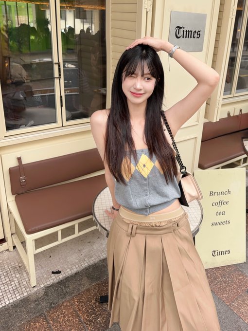 A young woman with long dark hair poses outside a cafe marked 'Times', wearing a gray sweater vest and tan pleated skirt, with a sign listing 'Brunch, coffee, tea, sweets'.