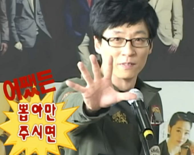 A photo of Korean comedian Yoo Jae-suk wearing glasses and a green jacket, holding up his hand toward the camera, with Korean text '어쨌든 범여만 주시면' (meaning 'Anyway, if you just give me a bonus').