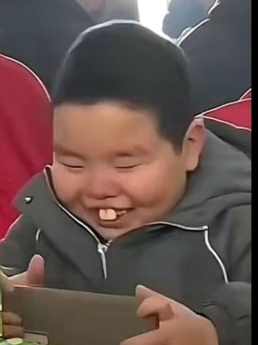 The image features a small boy with black hair sitting down, smiling while holding a cell phone up in front of his face. He seems happy about something he's doing or seeing on the screen. On the lower left corner of the photo, there is a caption that reads "大饼周线级别无停". The background appears to be grayish-brown, which complements the main subjects and adds context to the image's overall theme.