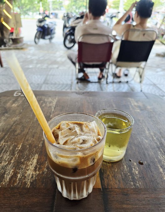 Two iced coffee drinks—one Vietnamese-style with condensed milk and a yellow straw, the other a green tea—sit on a wooden café table, with two people seated in chairs on a busy street with motorbikes visible in the background.