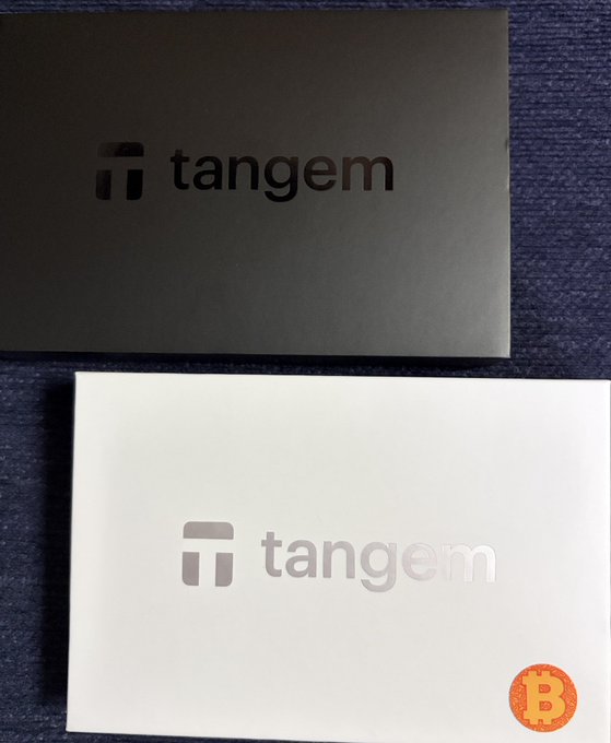 Two Tangem crypto hardware wallet boxes, one black and one white with a Bitcoin logo sticker, rest on a dark fabric surface.