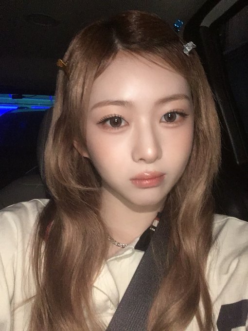 A young woman with long, wavy light brown hair and hair clips sits in a car at night, wearing a white top and a silver necklace with her seatbelt fastened across her chest.